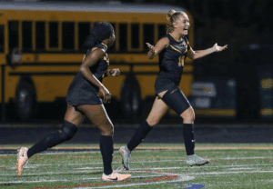 Two female soccer players celebrating on the field in front of a yellow school bus at night.