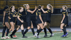 Girls high school soccer team players celebrating on field with high-fives and smiles in black uniforms.