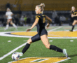 Female soccer player in black jersey kicking ball on field during night match.