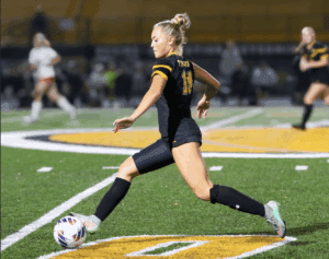 Female soccer player in black jersey kicking ball on field during night match.