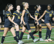 Women's soccer team in black jerseys celebrating on the field at night.