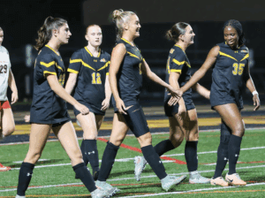 Women's soccer team in black jerseys celebrating on the field at night.