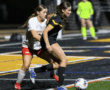 Girls' soccer match at night, player in black uniform dribbling past defender on a turf field.