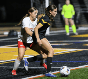 Girls' soccer match at night, player in black uniform dribbling past defender on a turf field.