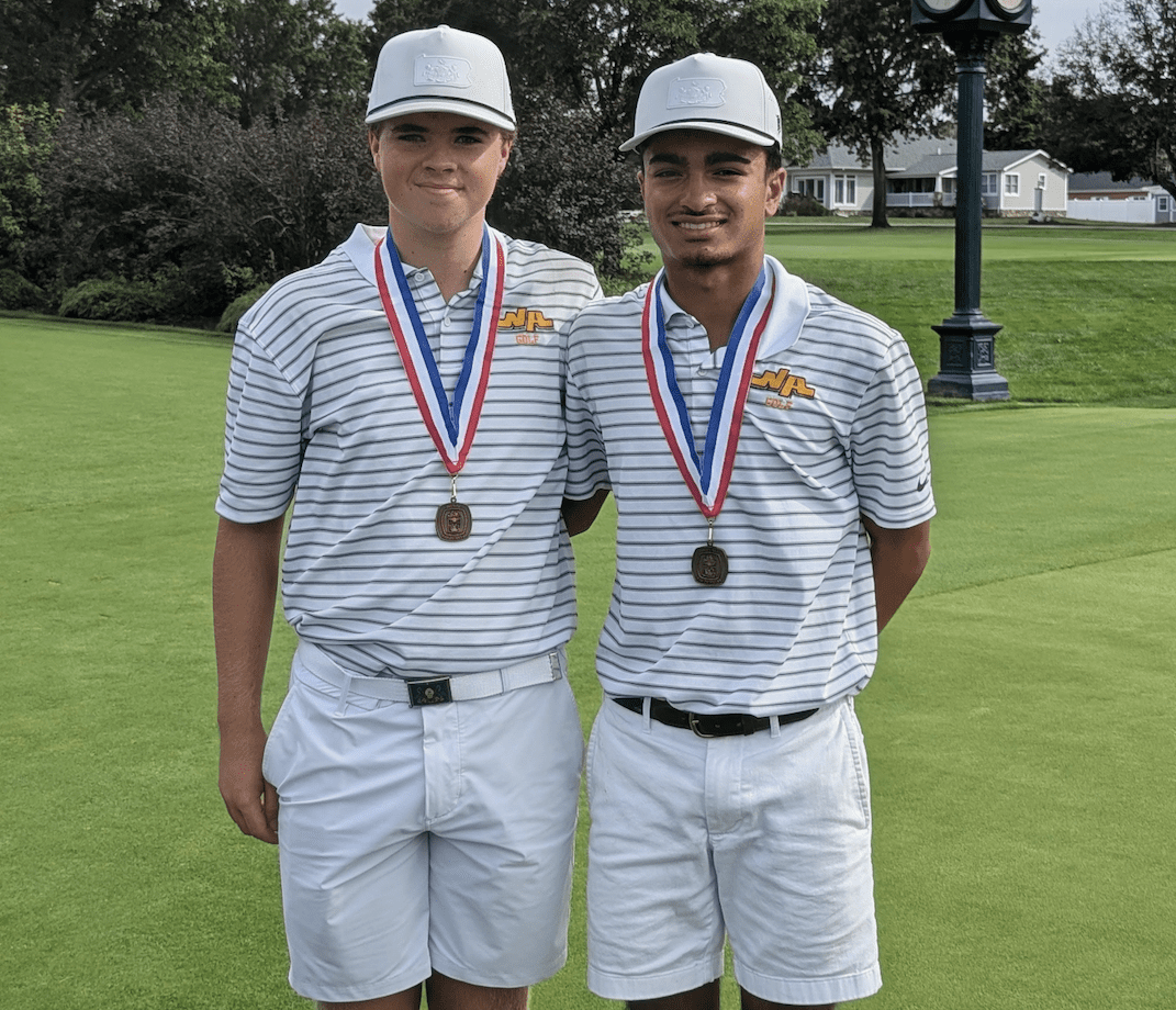 Two golfers wearing medals pose on a green field, showcasing their tournament success in matching outfits and caps.