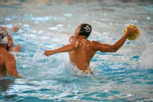 Water polo player in black cap prepares to throw ball in mid-action pool scene, splashing water everywhere.