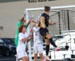 Soccer players leap to head ball near goalpost in intense match action.
