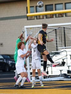 Soccer players leap to head ball near goalpost in intense match action.