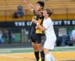 Two soccer players jump for the ball during an intense match on a field with yellow bleachers.