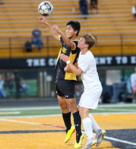 Two soccer players jump for the ball during an intense match on a field with yellow bleachers.
