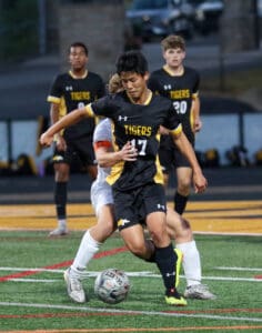Soccer players competing for the ball during a match, one in a black Tigers jersey, on a green field.