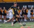 Soccer player in black and yellow kit controls the ball during a match, with cheerleaders in the background.