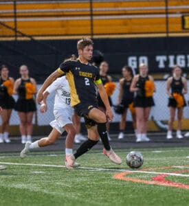 Soccer player in black and yellow kit controls the ball during a match, with cheerleaders in the background.
