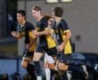 Soccer players in black and yellow uniforms running on the field during a game at dusk.