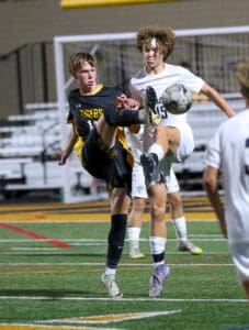 Soccer players in action during a night match, both leaping to kick the ball near the goal.