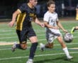 Two high school soccer players compete for the ball on a field during a night match.