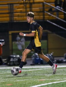 Soccer player in black and yellow kit dribbling on field under stadium lights.