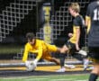 Soccer goalkeeper in yellow diving to save a ball during a night match on a turf field.