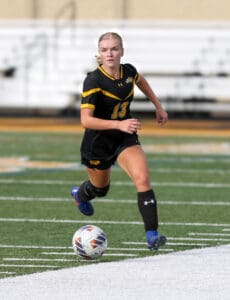 Soccer player in black and yellow uniform dribbles the ball on a green field during a game.