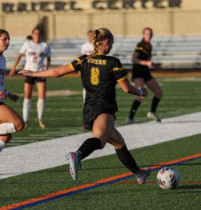 Soccer player in black jersey dribbles ball on field with opponents and teammates nearby.