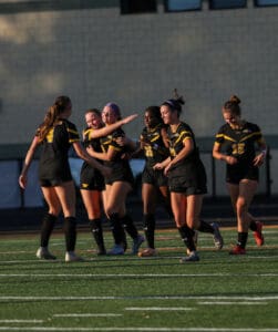 Soccer team celebrating on the field in black and yellow uniforms after a successful play, showing teamwork and joy.