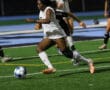 Soccer player dribbles ball during night game on field, wearing white uniform and knee brace.