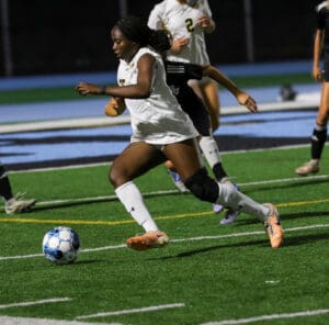 Soccer player dribbles ball during night game on field, wearing white uniform and knee brace.