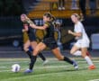 Soccer player in black uniform dribbles the ball past opponent during a night match on a field.