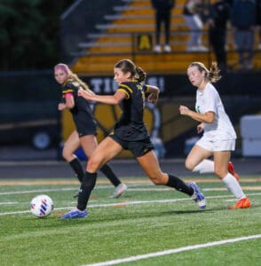 Soccer player in black uniform dribbles the ball past opponent during a night match on a field.