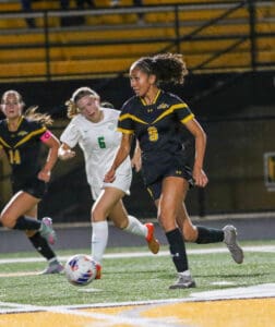 Soccer player in black and yellow uniform dribbles ball on field during a match with opposing team players nearby.