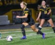 Female soccer player in black uniform dribbling ball on the field during a night game.