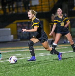 Female soccer player in black uniform dribbling ball on the field during a night game.