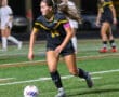 Female soccer player in black uniform dribbles ball during a match on a green field, focused on strategy.