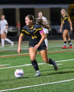 Female soccer player in black uniform dribbles ball during a match on a green field, focused on strategy.