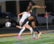 Two female soccer players compete for the ball in an intense night match on a well-lit field.
