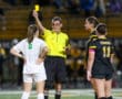Referee issues yellow card to soccer player during a game, with players in white and black uniforms watching.