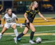 Two female soccer players compete for possession during a night game on a field with school buses in the background.