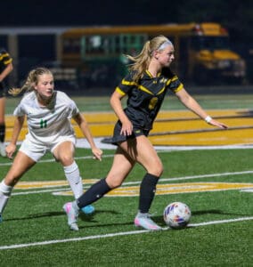 Two female soccer players compete for possession during a night game on a field with school buses in the background.