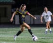 Soccer player in black uniform dribbles ball during night match on grass field, opponent in white follows closely.