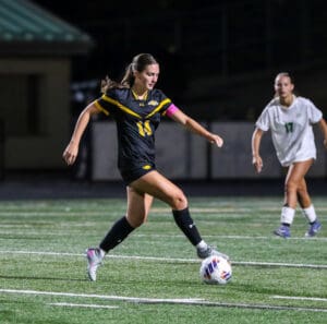 Soccer player in black uniform dribbles ball during night match on grass field, opponent in white follows closely.