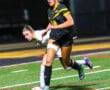 Soccer player in black uniform dribbles ball on field during night game.