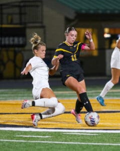 Soccer players in action during a nighttime game on a field, one wearing black and gold, the other in white.