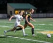 Two female soccer players racing towards a soccer ball on a field at night.