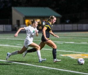 Two female soccer players racing towards a soccer ball on a field at night.