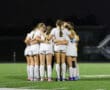 Girls' soccer team huddles on the field at night, showcasing unity and teamwork before a match.