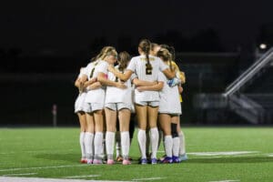 Girls' soccer team huddles on the field at night, showcasing unity and teamwork before a match.
