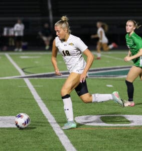 Soccer player in white jersey dribbles ball on field during night match, followed by opponent in green.
