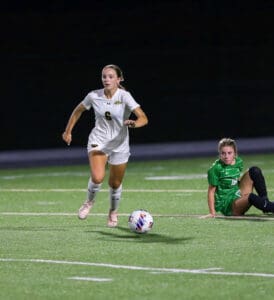 Soccer player in white jersey dribbles ball on field, opponent in green on the ground, intense match action.