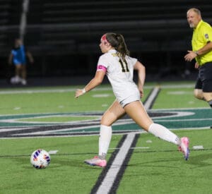 Soccer player in white dribbling ball on field, wearing captain's armband, with referee nearby during night match.