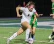 Soccer player in white jersey dribbles past defender during nighttime match on grass field.